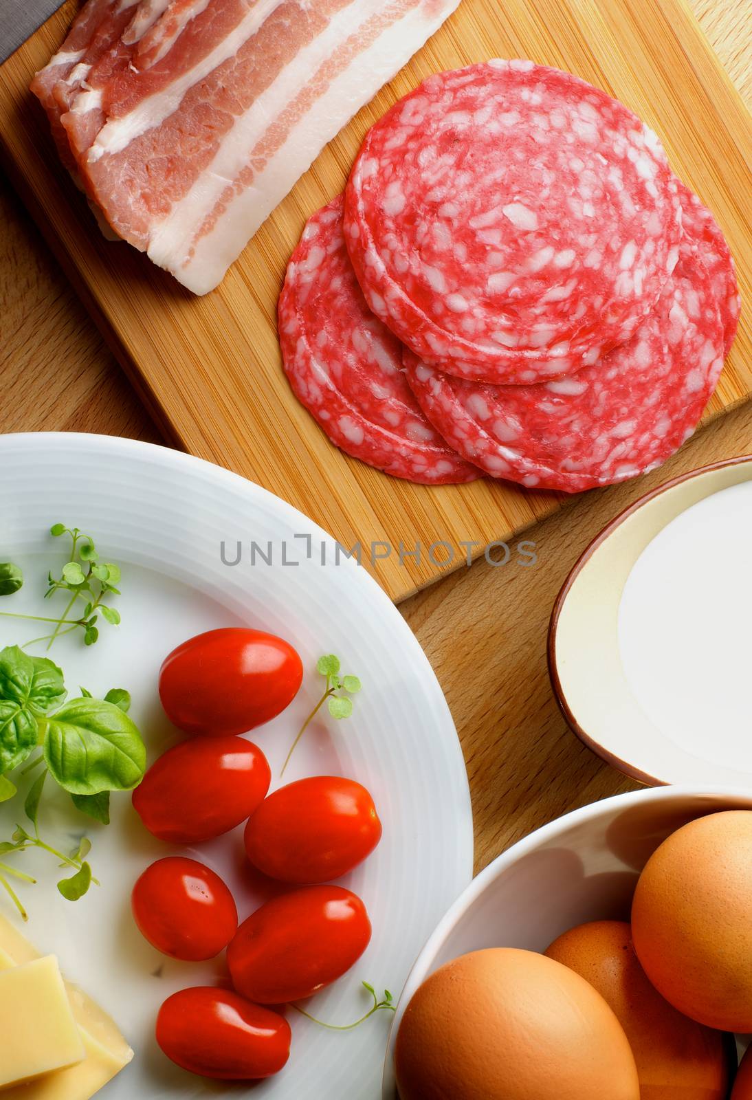 Ingredients of Omelet with Eggs, Milk, Cherry Tomatoes, Sausage, Bacon and Greens in Bowls on Wooden Cutting Board closeup