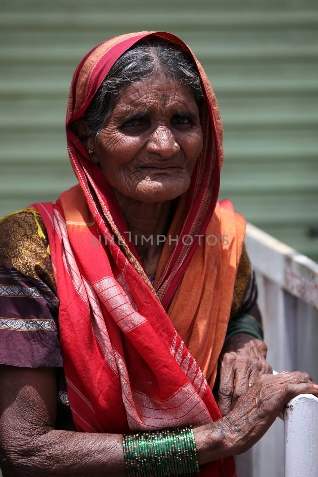 A very old female pilgrim from India standing against in the sun holding the police barricades, in India.