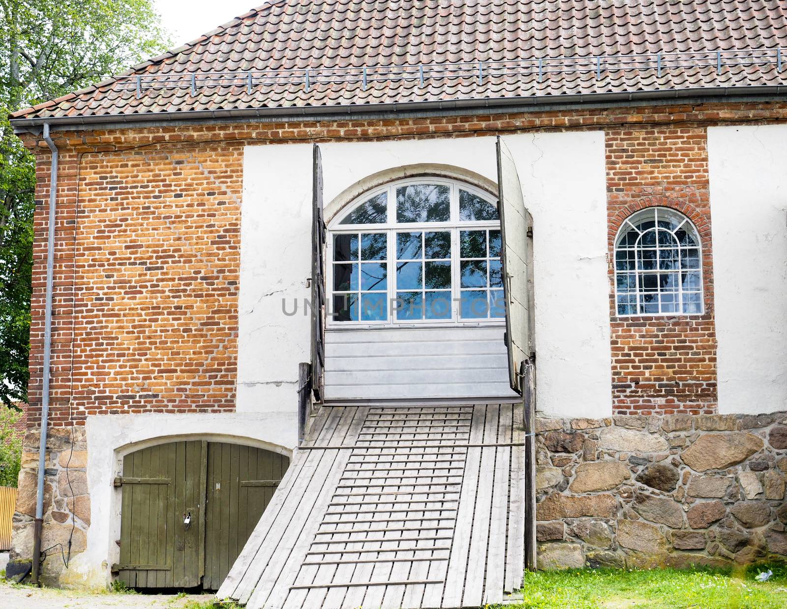 Old red brick building with wooden bridge path towards blue windows, dark roof stones