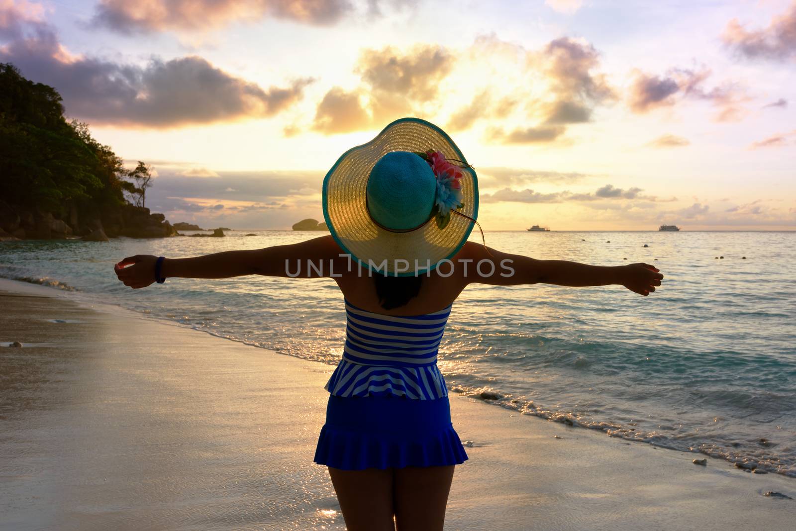 Tourist girl in a swimsuit blue and white striped hat standing on beach with pleasure the beautiful landscape of sky and sea during sunrise, Miang island, Similan National Park, Phang Nga, Thailand