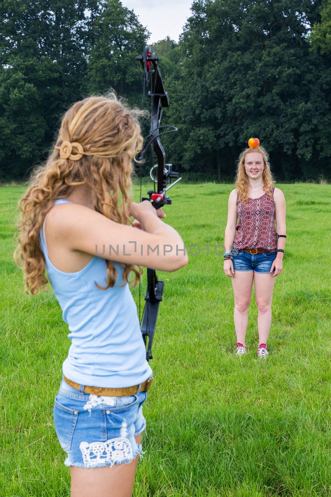 Teenage girl aiming arrow of compound bow at apple on head of young woman