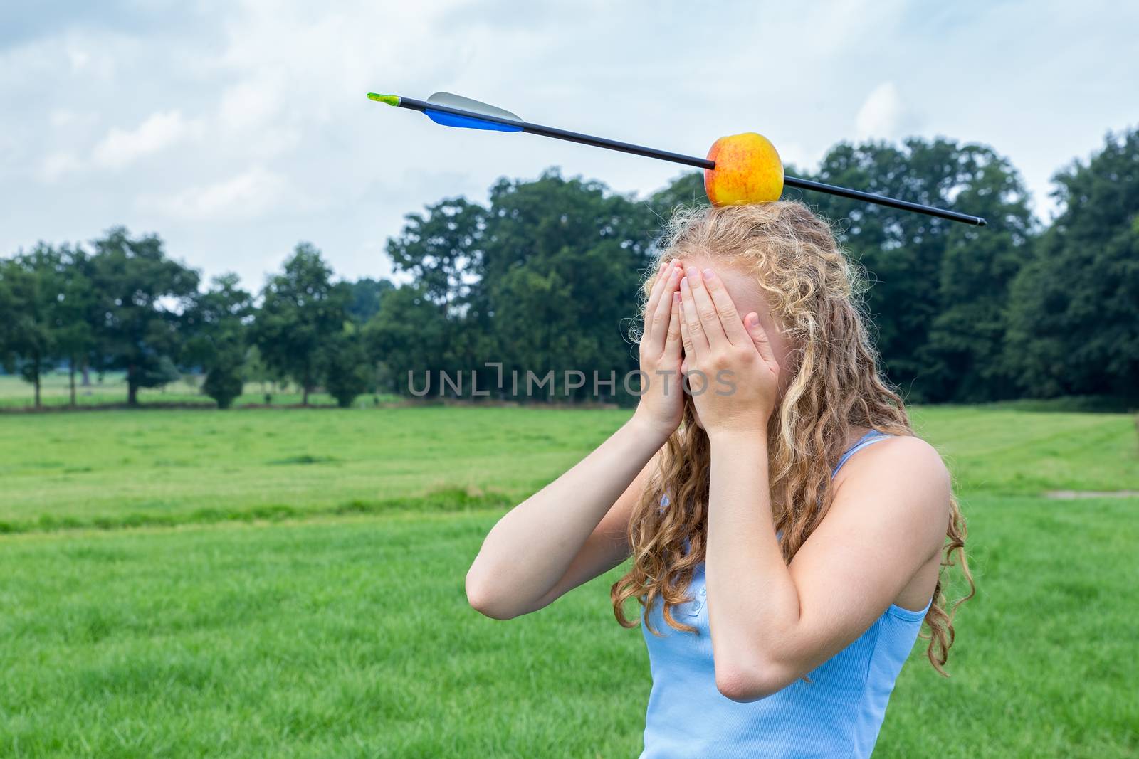 Teenage girl covering her face feeling fearful with apple and arrow on head in green meadow outdoors