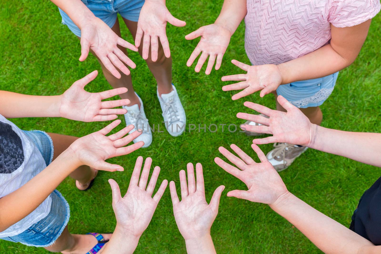 Ten arms of children in circle with palms of hands up above green grass