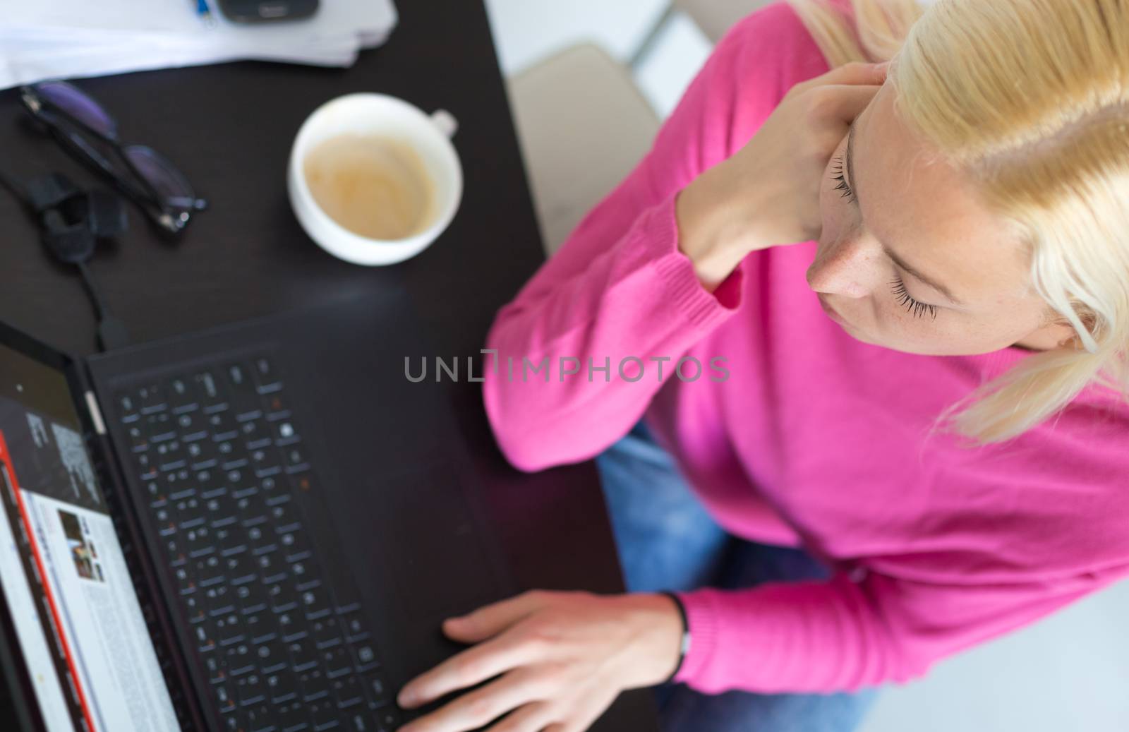 Female freelancer in her casual home clothing remotly working on laptop computer from her home while drinking her morning cup coffee. View from above.