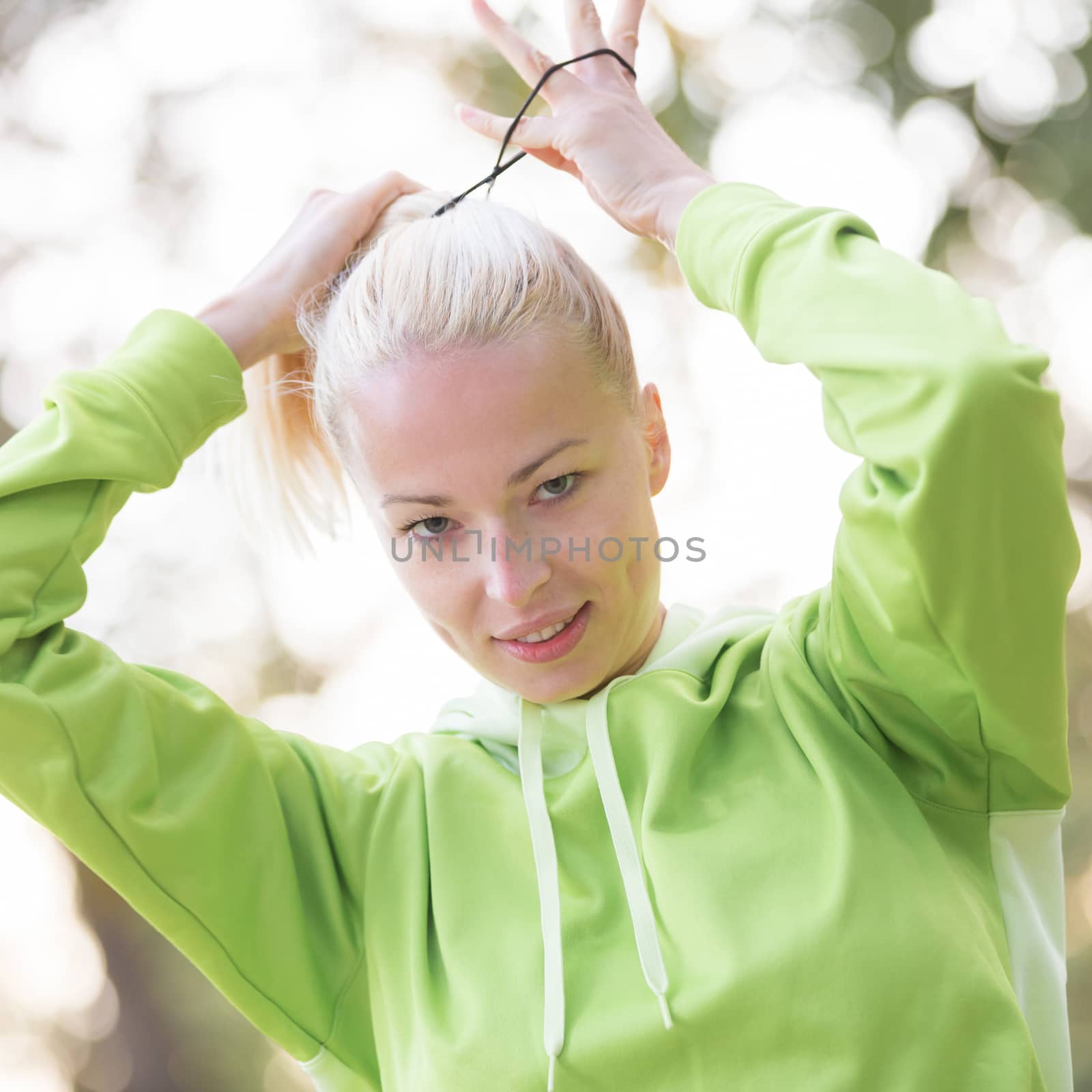 Portrait of attractive confident sporty woman wearing fashionable green sportswear  making her hair in ponytail before training in nature. Active sporty lifestyle concept.