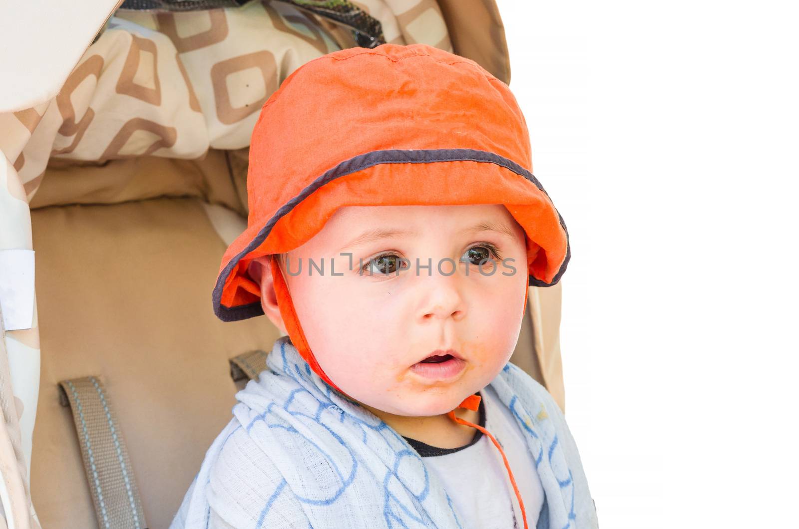 Baby with orange cap sitting in a stroller, looking amazed. Background isolated.