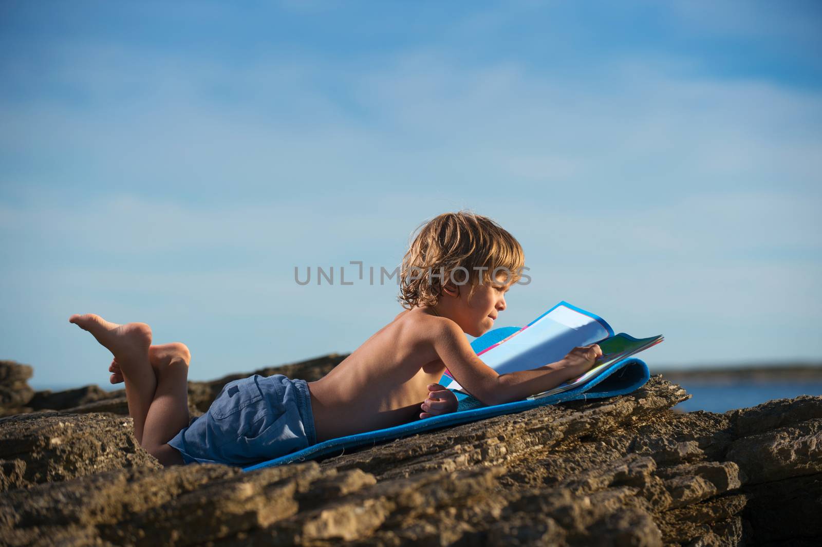 Cute little boy lying on his stomach looking at the picture book on the beach. Somen negative space around.