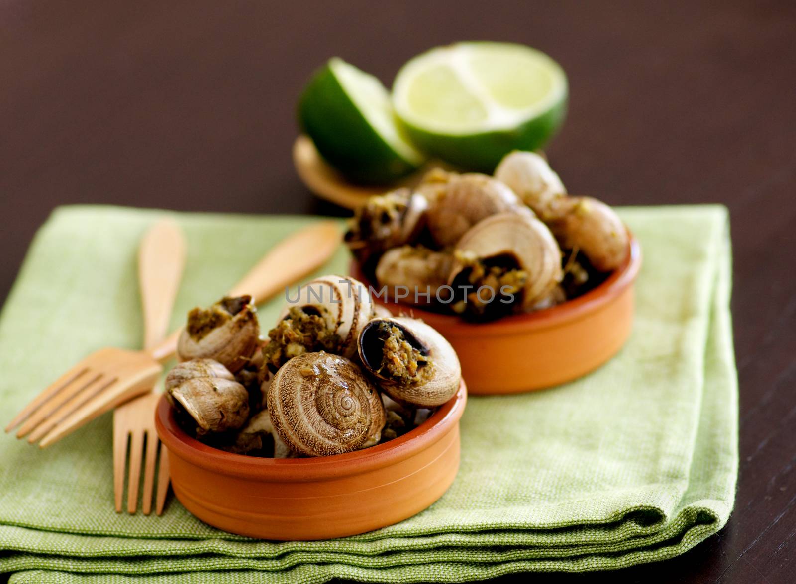 Delicious Escargot with Garlic Butter in Two Bowls with Wooden Forks and Halves of Lime closeup on Green Napkin. Focus on Foreground