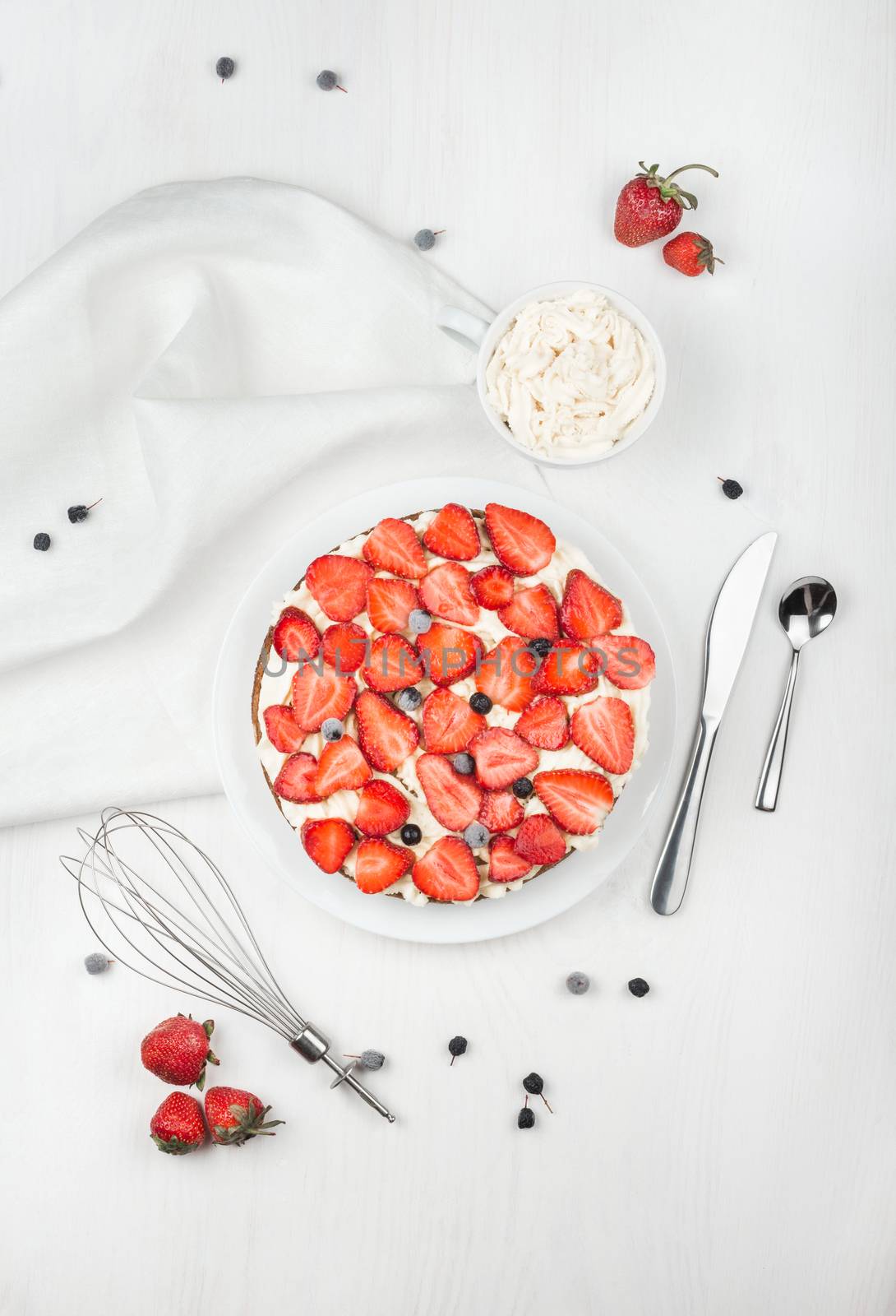 Delicious nutritious cake with fresh strawberries decorated with chokeberry, white cup with whipped cream, steel spoon, whisk, strawberry, napkin, plate, top view,  good morning