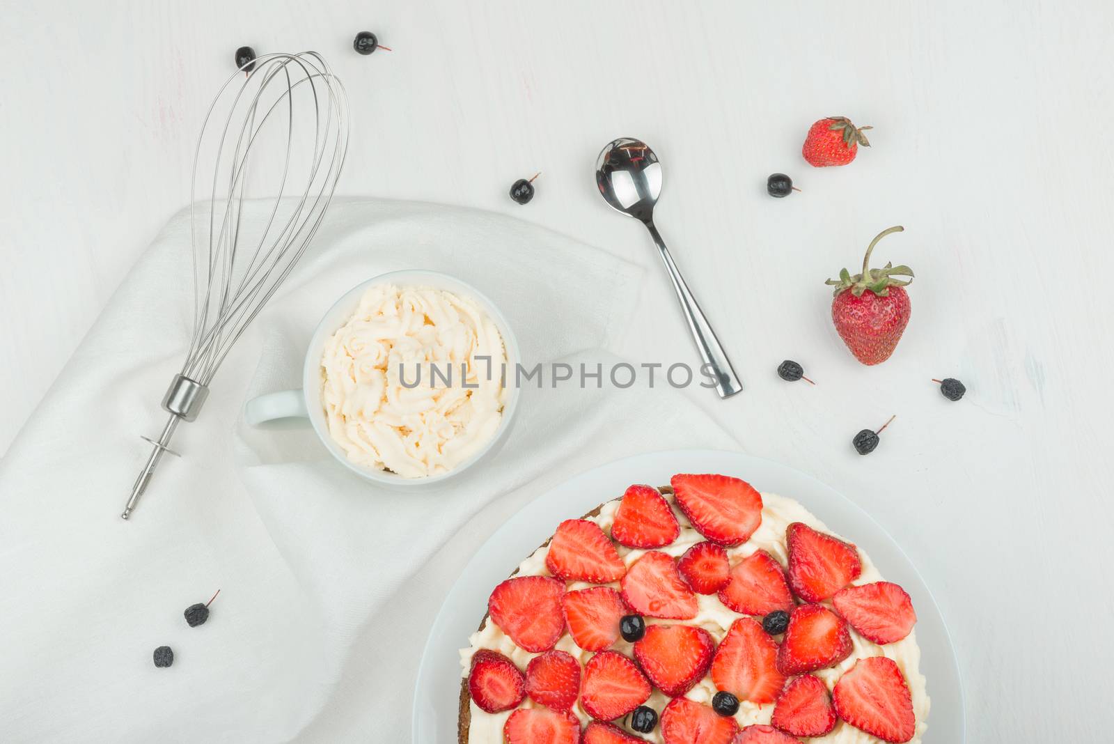 Delicious nutritious cake with fresh strawberries decorated with chokeberry, white cup with whipped cream, steel spoon, whisk, strawberry, napkin, plate, top view,  good morning