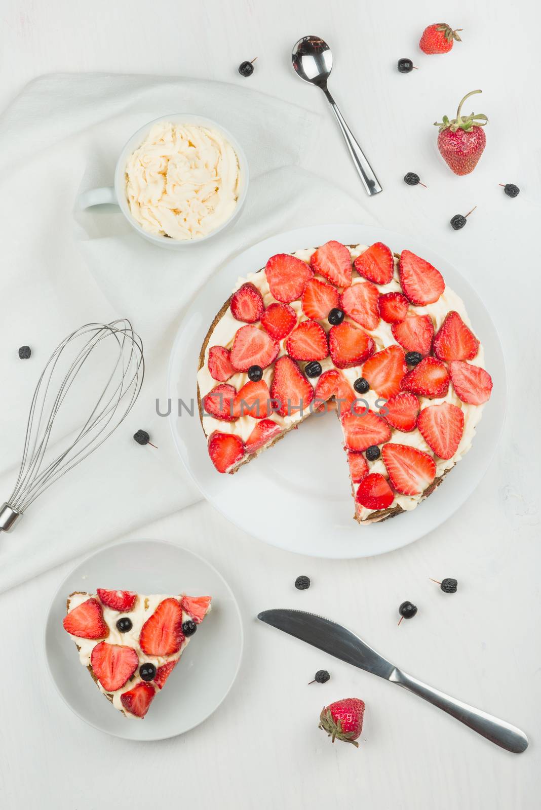 Delicious nutritious cake with fresh strawberries decorated with chokeberry, white cup with whipped cream, steel spoon, whisk, strawberry, napkin, plate, top view,  good morning