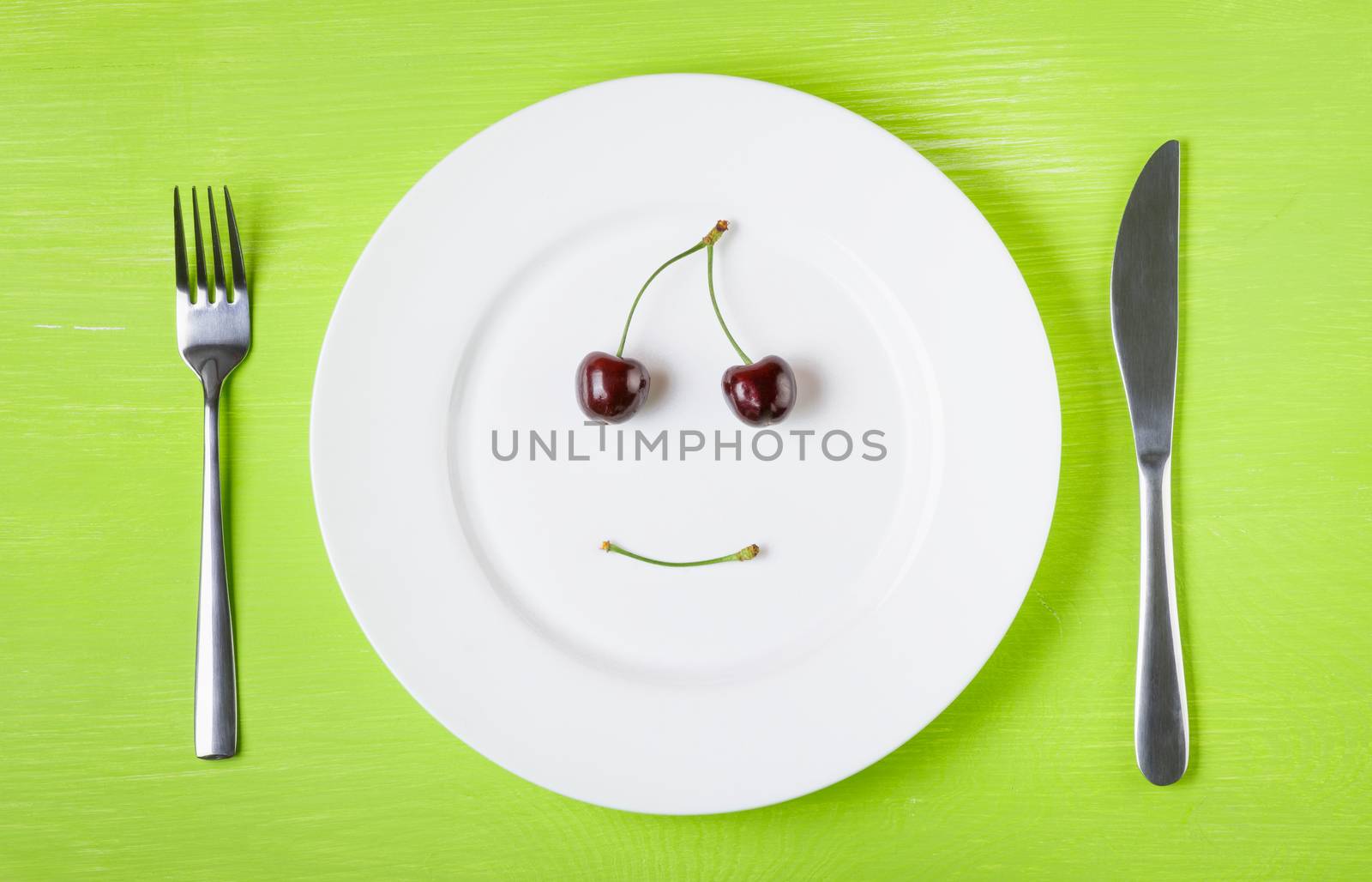 Smiling face of cherries on a white plate, knife and fork on a bright green table. The concept of a raw food diet, vegetarian, healthy eating, diet, weight loss, good mood