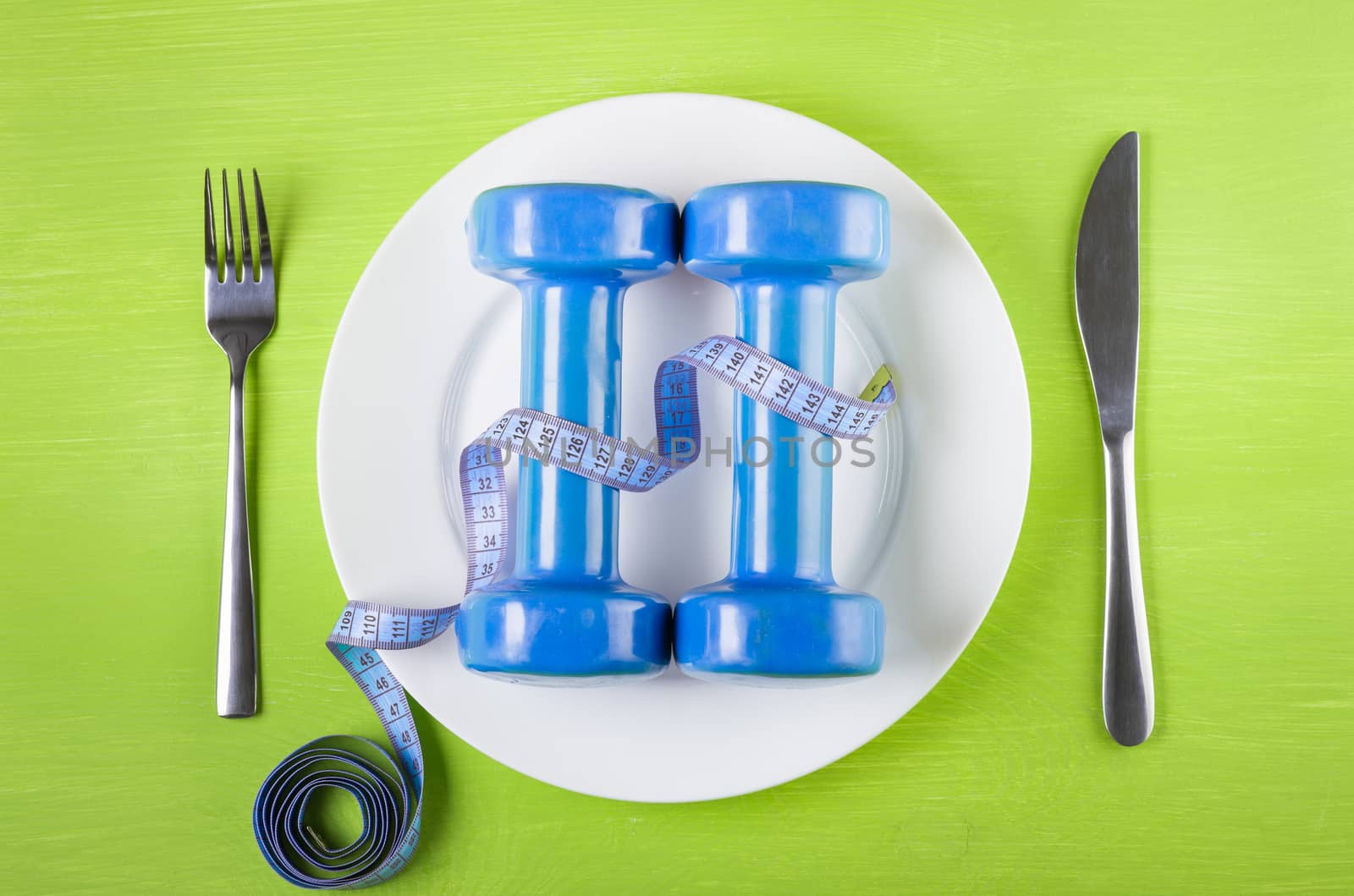 The concept of a healthy lifestyle, diet, sports, weight loss, anti-obesity, exercise, healthy diet.  Centimeter abd dumbbells on a plate, knife  and fork on a green table, top view, closeup