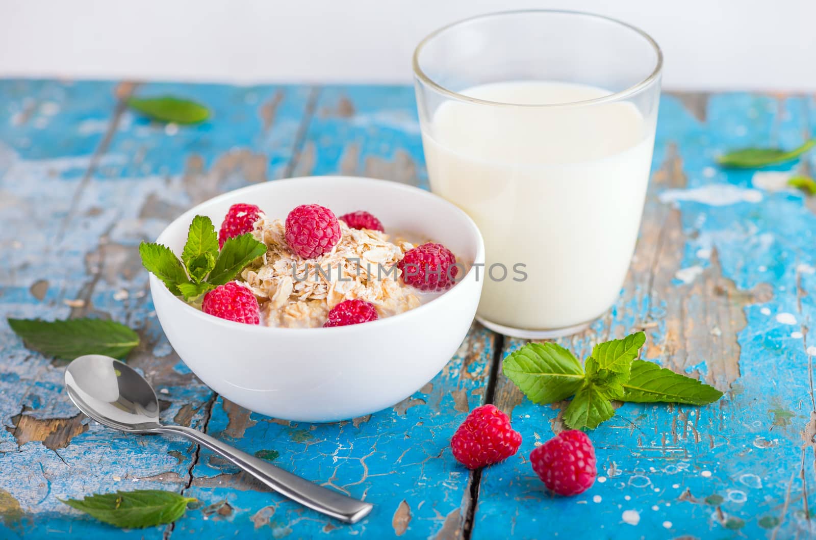 Oat flakes with milk and frash raspberries for breakfast, glass with milk, spoon, fresh mint on an old wooden blue background. The concept of a healthy diet, weight loss