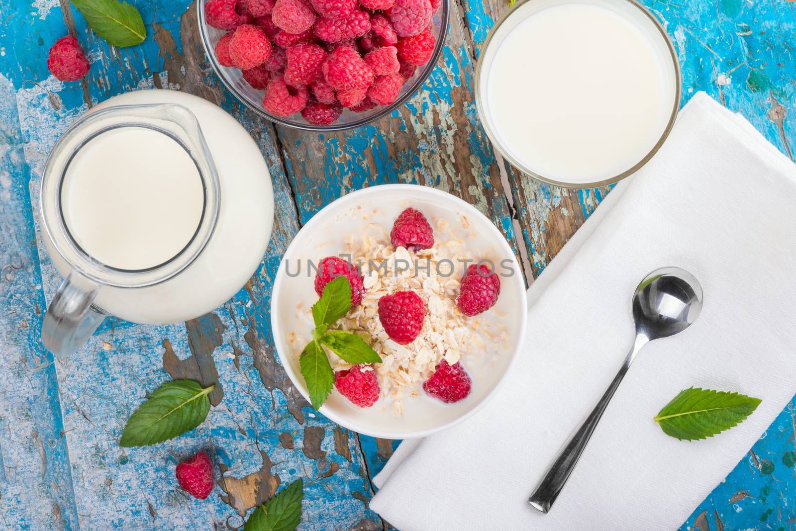 Oat flakes with milk and raspberries for breakfast Jug and glass with milk, spoon, napkin, fresh mint on an old wooden blue background, top view. The concept of a healthy diet, weight loss