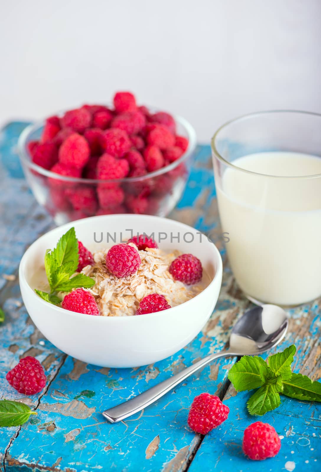 Oat flakes with milk and frash raspberries for breakfast, glass with milk, spoon, fresh mint on an old wooden blue background. The concept of a healthy diet, weight loss