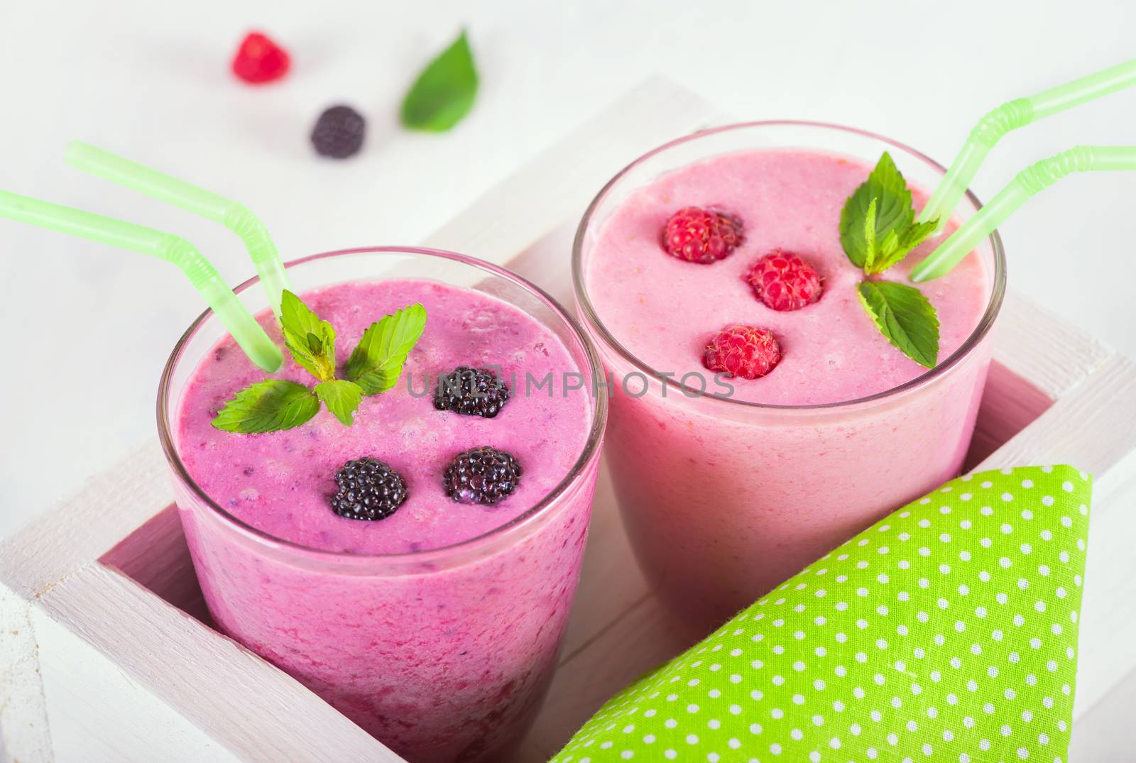 Raspberry and blackberry dairy smoothies with fresh berries and sprigs of mint, green cocktail stick in a decorative white box on a white wooden table