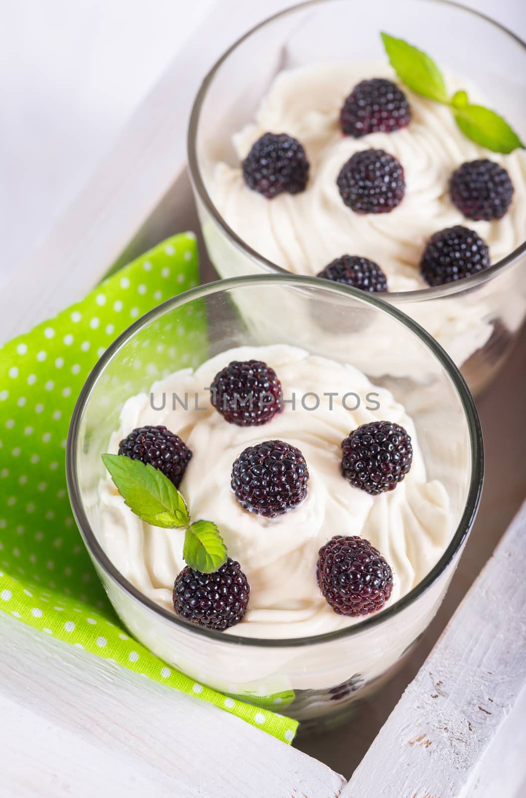 delicious soft cheese dessert with whipped cream and fresh blackberries with a sprig of fresh mint in a glass, green napkin in a decorative wooden box on a white table