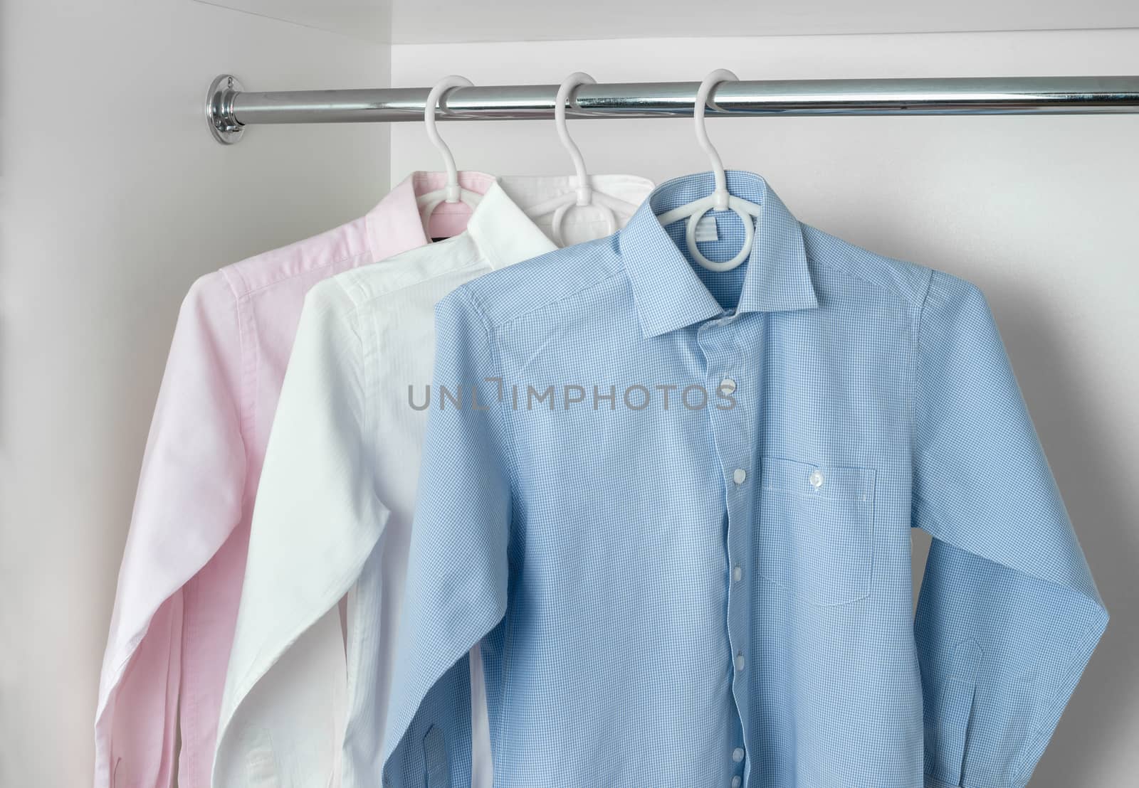 white, blue and pink clean ironed men's shirts hanging on hangers in the white wardrobe