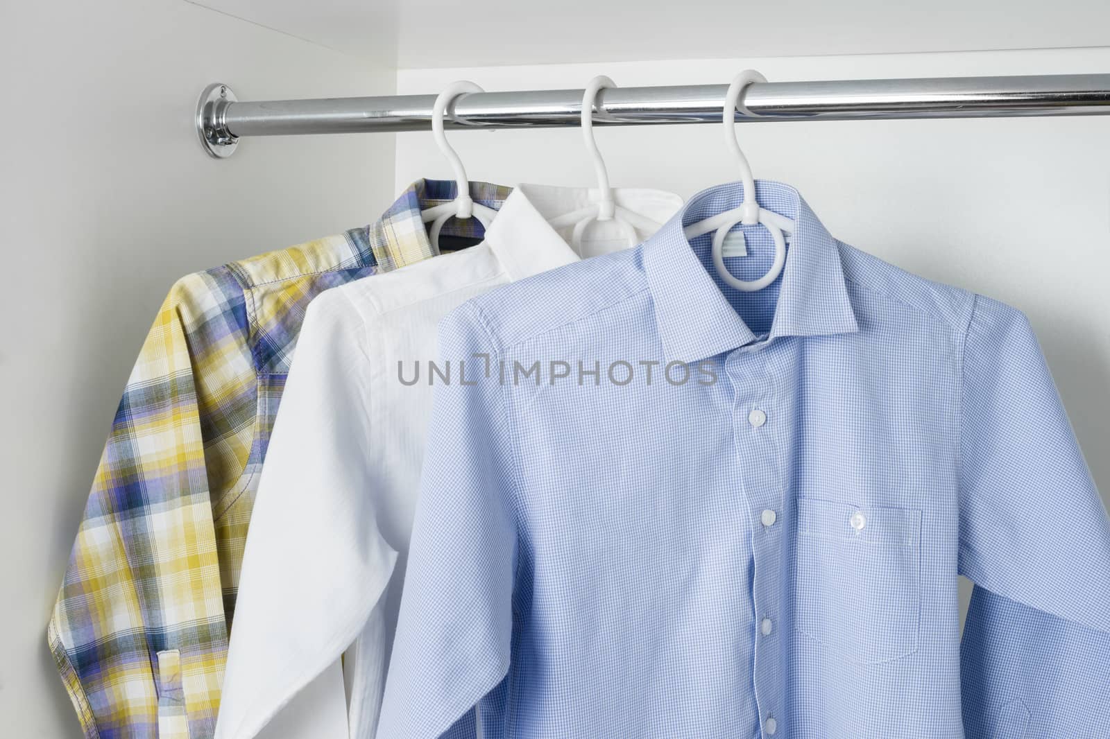 white, blue and checkered clean ironed men's shirts hanging on hangers in the white wardrobe 