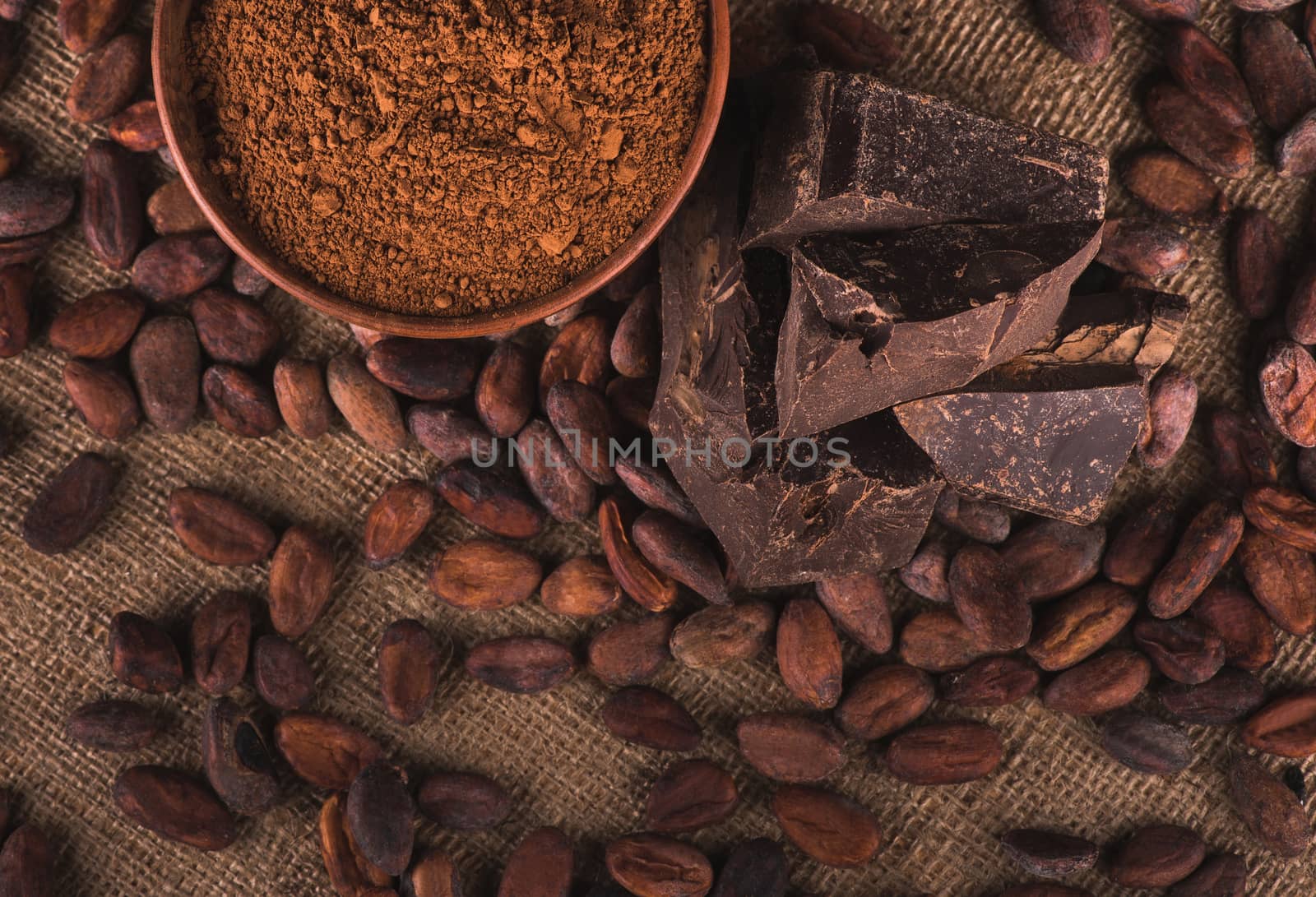 Raw cocoa beans, clay bowl with cocoa powder, black chocolate on brown sacking, top view