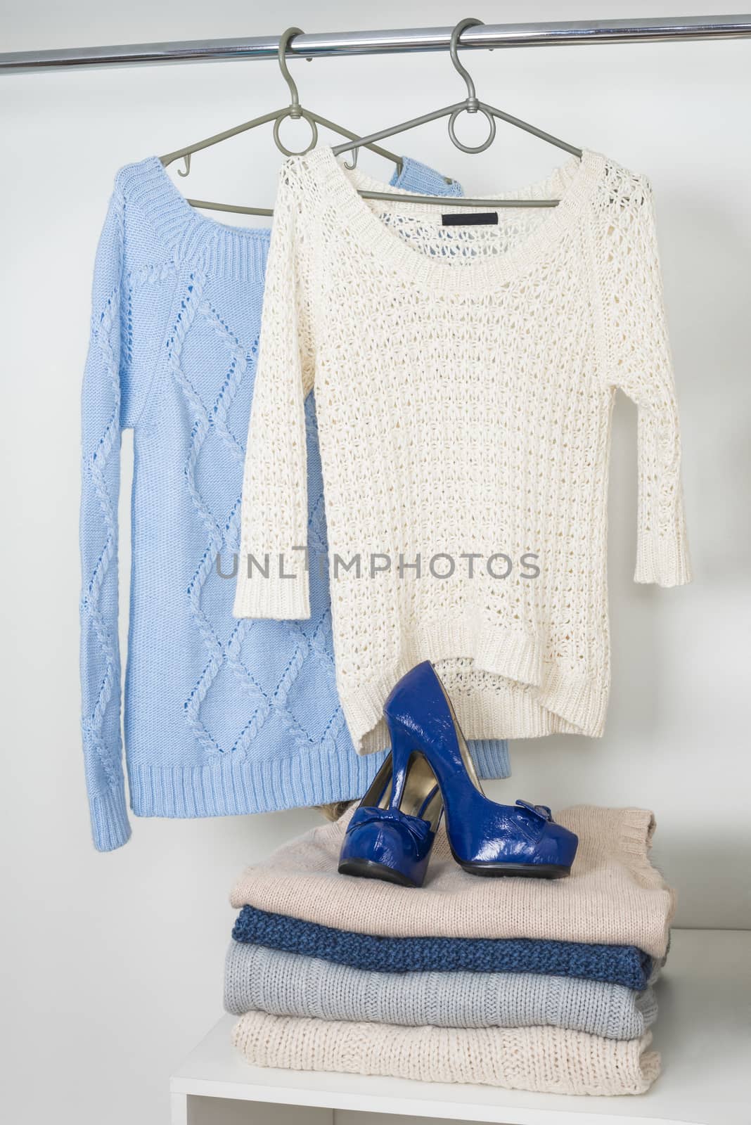 Women's warm knitted things in the white cupboard, two sweaters hanging on hangers, a pile of warm woolen clothes and lacquered blue shoes
