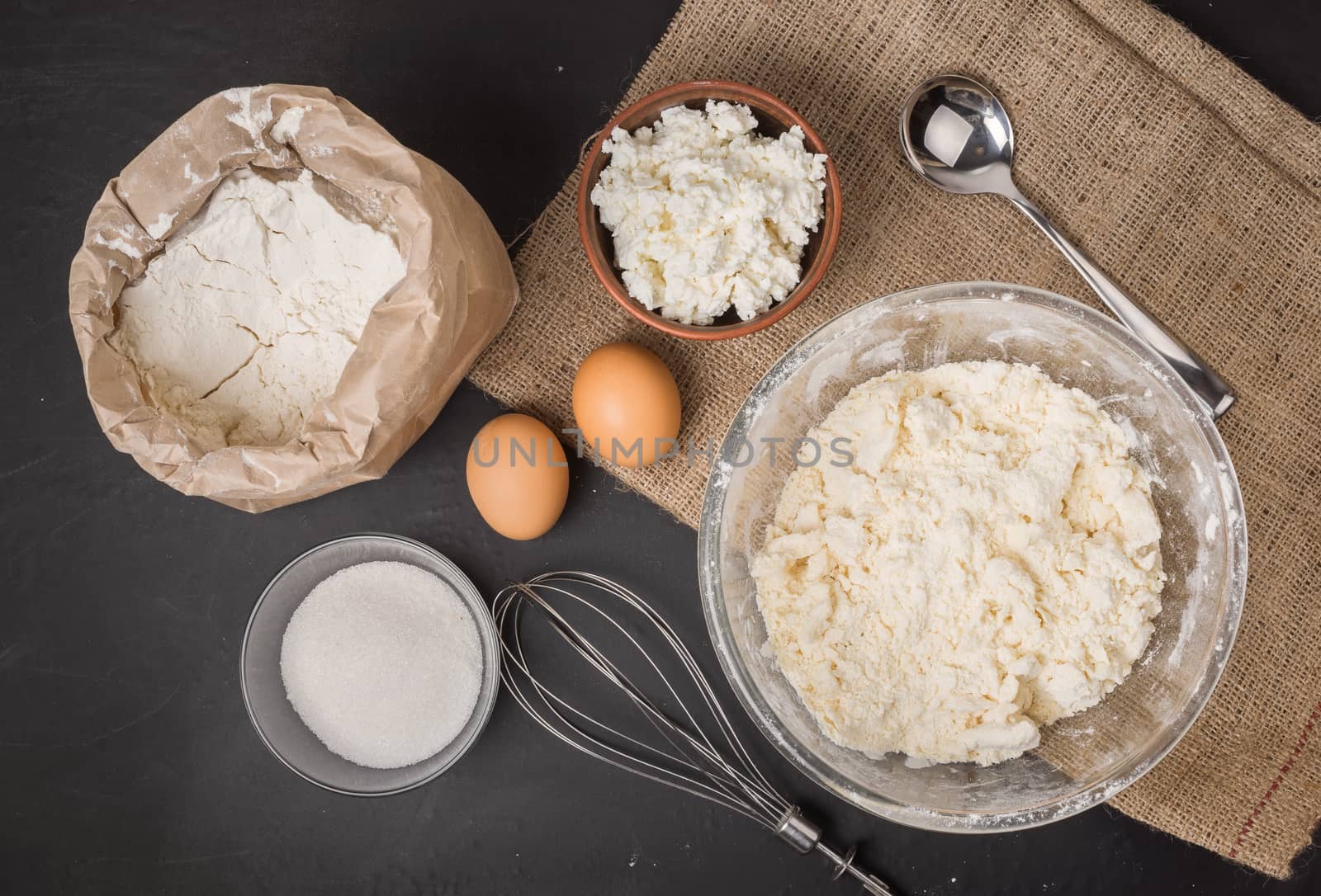 The ingredients for homemade baking cheesecake, a wheat flour in a paper bag, two eggs, sugar in a bowl, whisk and a spoon on sackcloth, top view