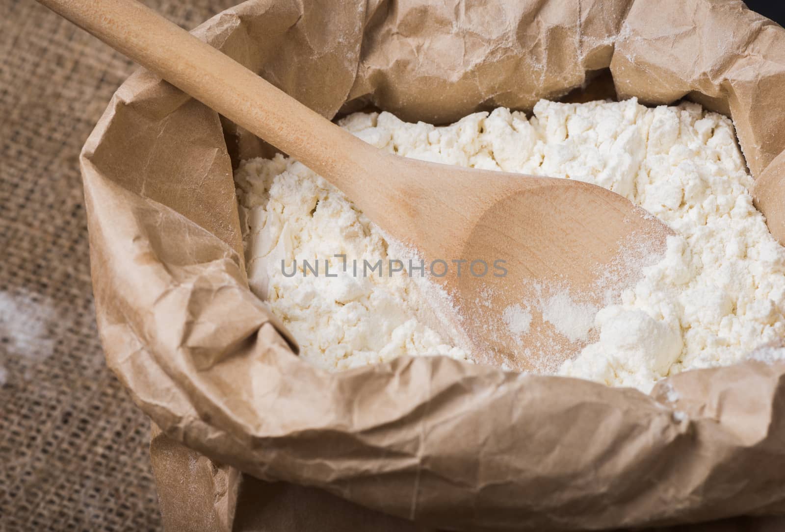 Whole flour in a paper bag and a wooden spoon on sacking, close up