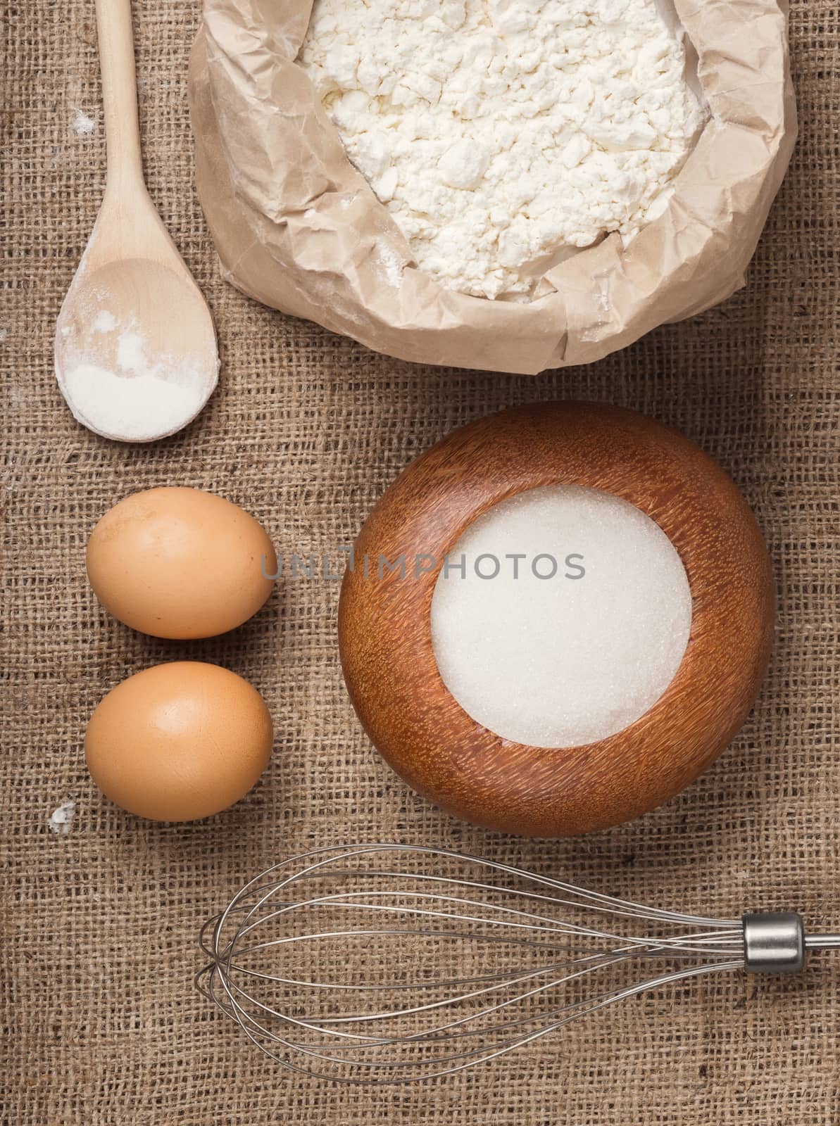 The ingredients for homemade baking sponge cake, a wheat flour in a paper bag, two eggs, sugar in a wooden bowl, whisk and a wooden spoon on sackcloth, top view