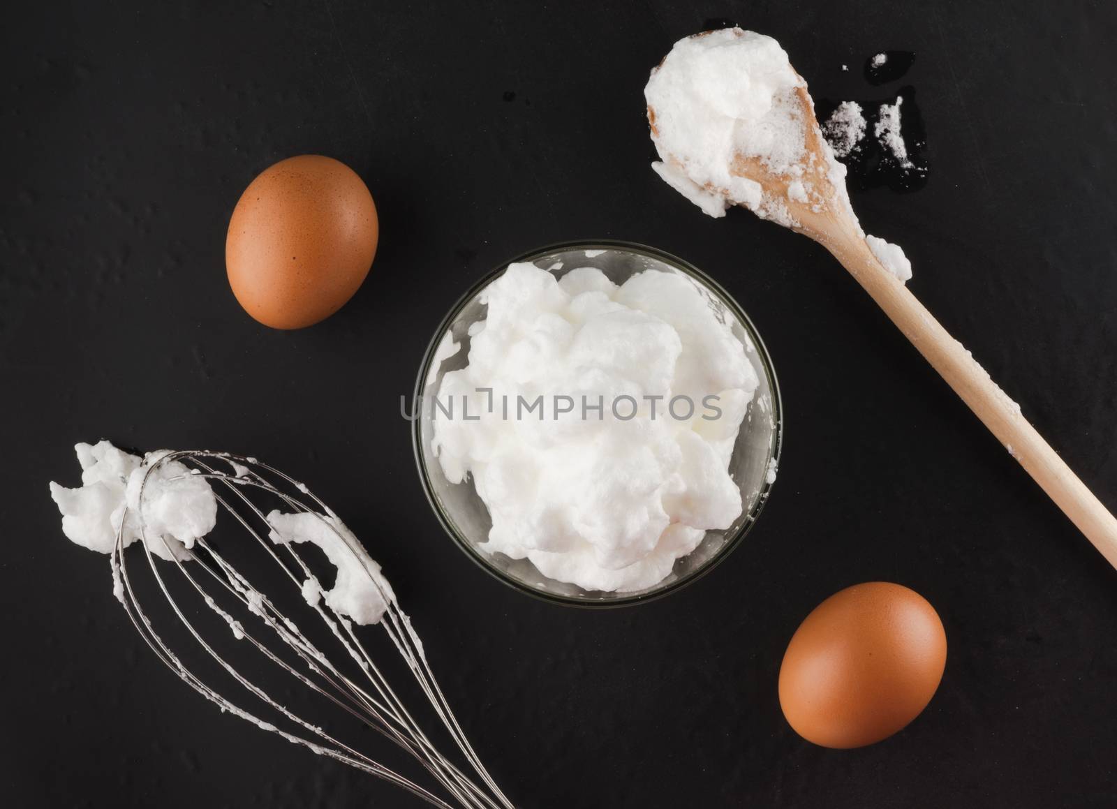 Whipped egg whites for cream in a glass bowl, whisk and wooden spoon on a dark background, top view