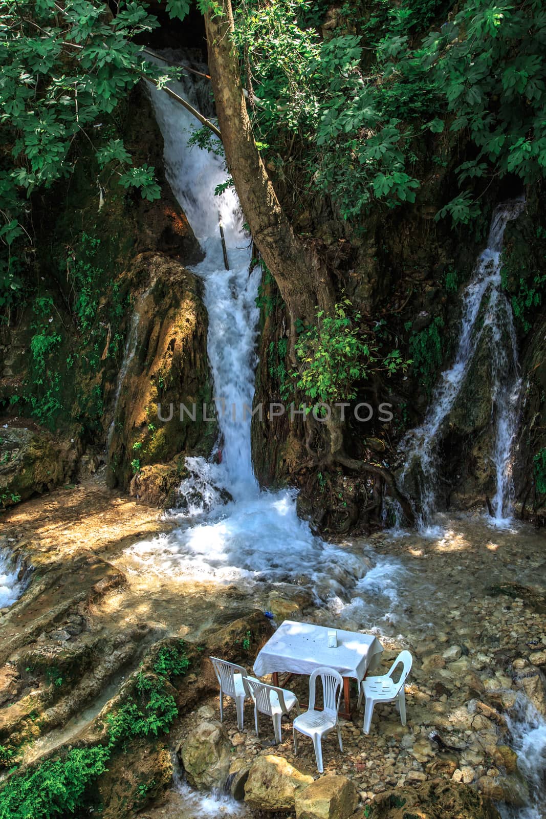 View of the tables and chairs near the flowing water of Harbiye Waterfall on natural rocks among trees.