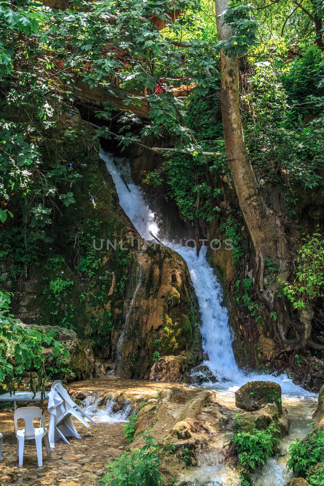 View of the tables and chairs near the flowing water of Harbiye Waterfall on natural rocks among trees.