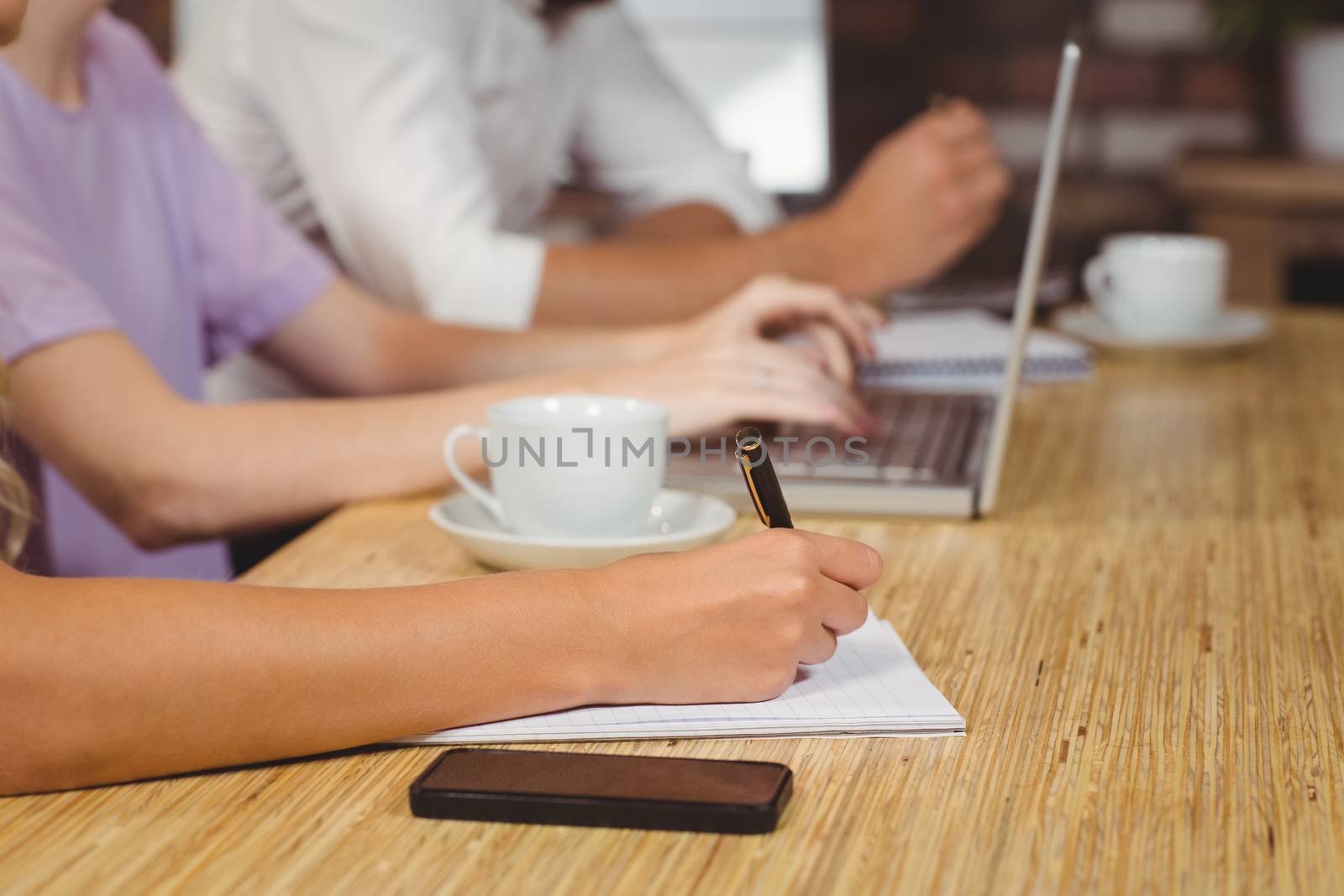 Woman writing on book with colleagues working on technologies in background 