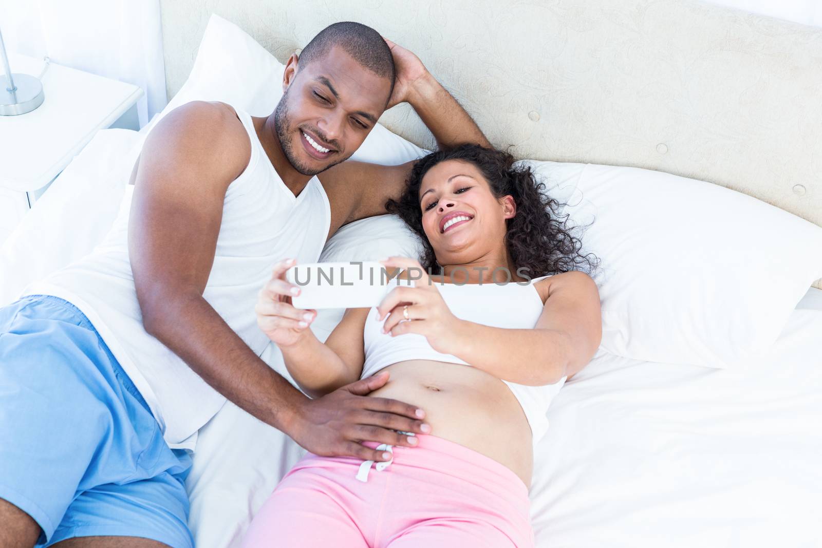 Pregnant wife showing smartphone to husband lying on bed at home