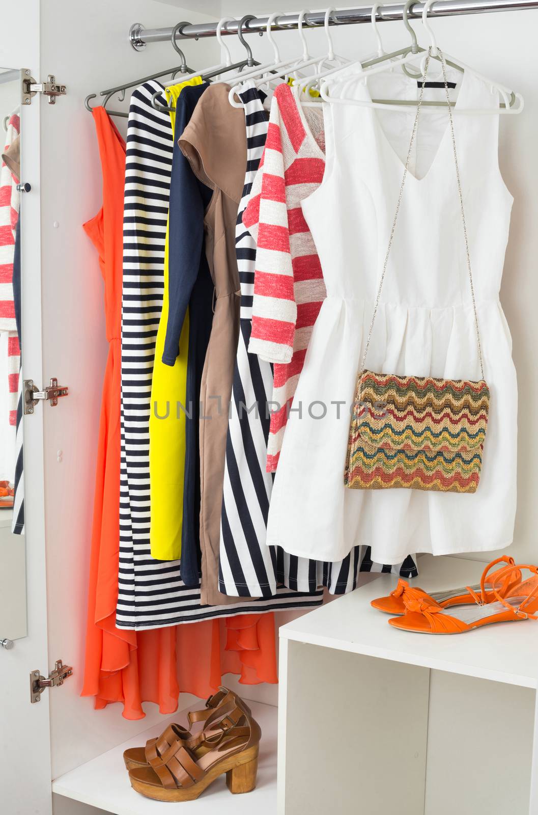 Set of bright colorful female fashion clothes on the hangers for spring and summer wardrobe, handbag, sandals in white cabinet, vertical