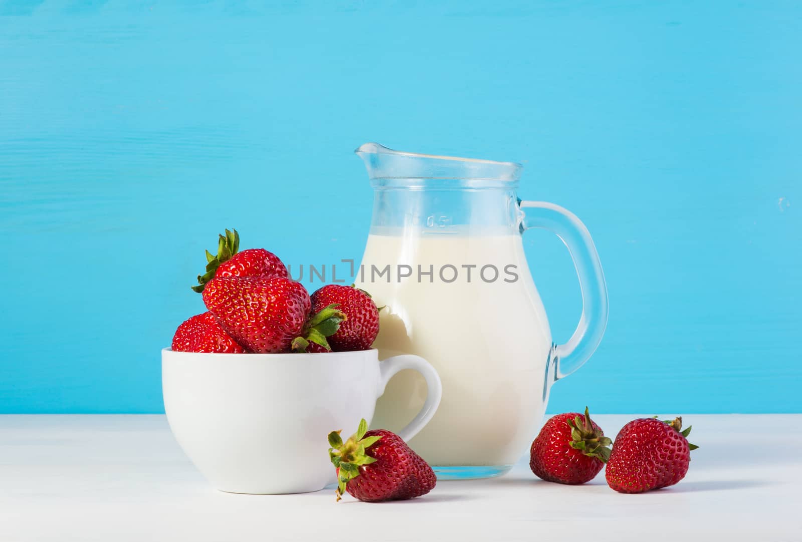 ingredients for strawberry milkshake: rustic fresh milk in a glass jar and raw ripe fresh strawberries in a white cup on a white wooden table on a blue background