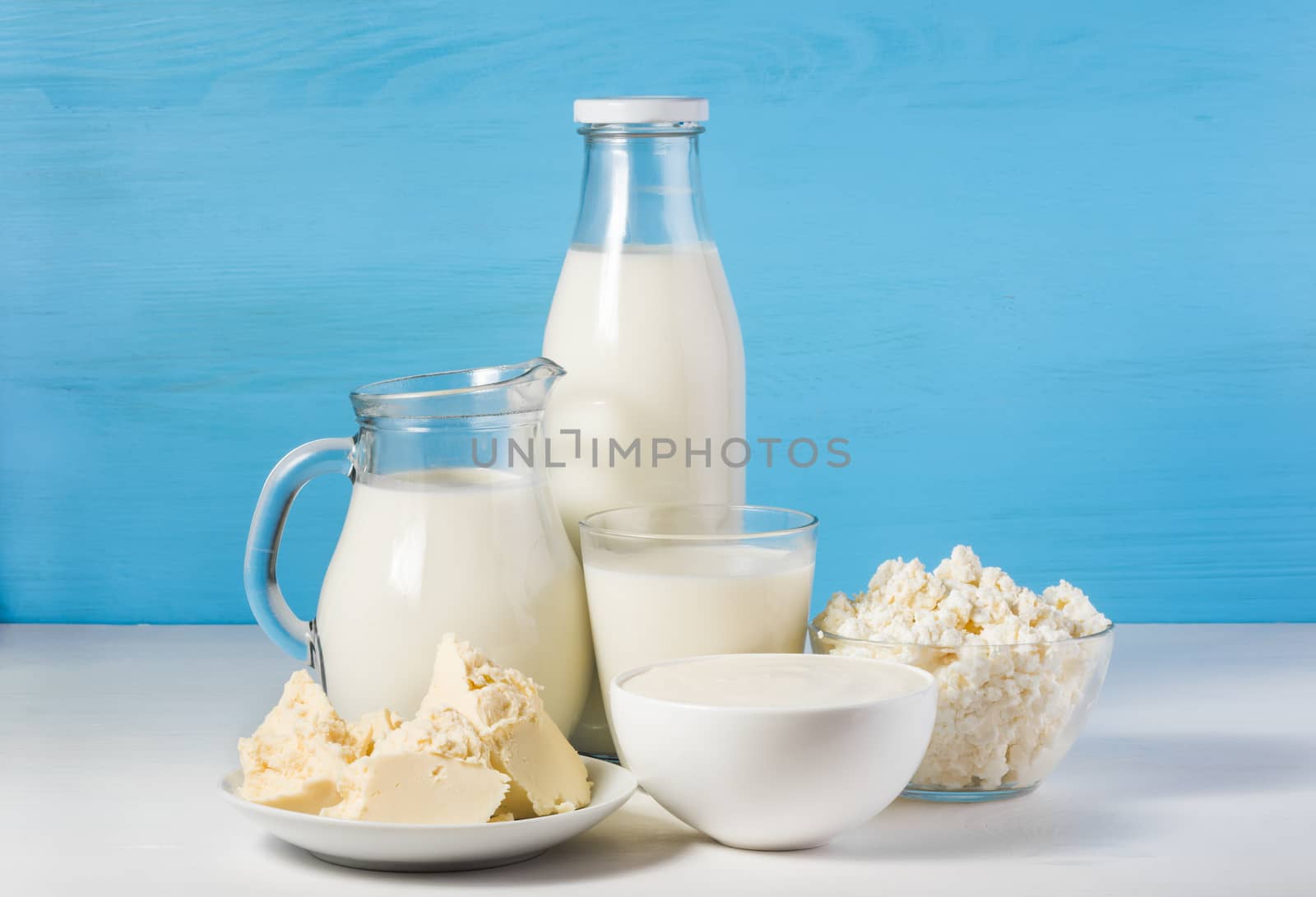 tasty healthy dairy products on a white table on a blue background: sour cream in a white bowl, cottage cheese in bowl, butter on a saucer and milk in a jar, glass bottle and in a glass