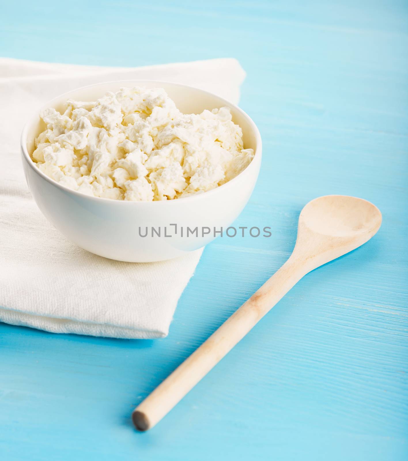 Tasty healthy village cottage cheese in a white ceramic bowl, a white kitchen cloth and wooden spoon on a blue wooden table