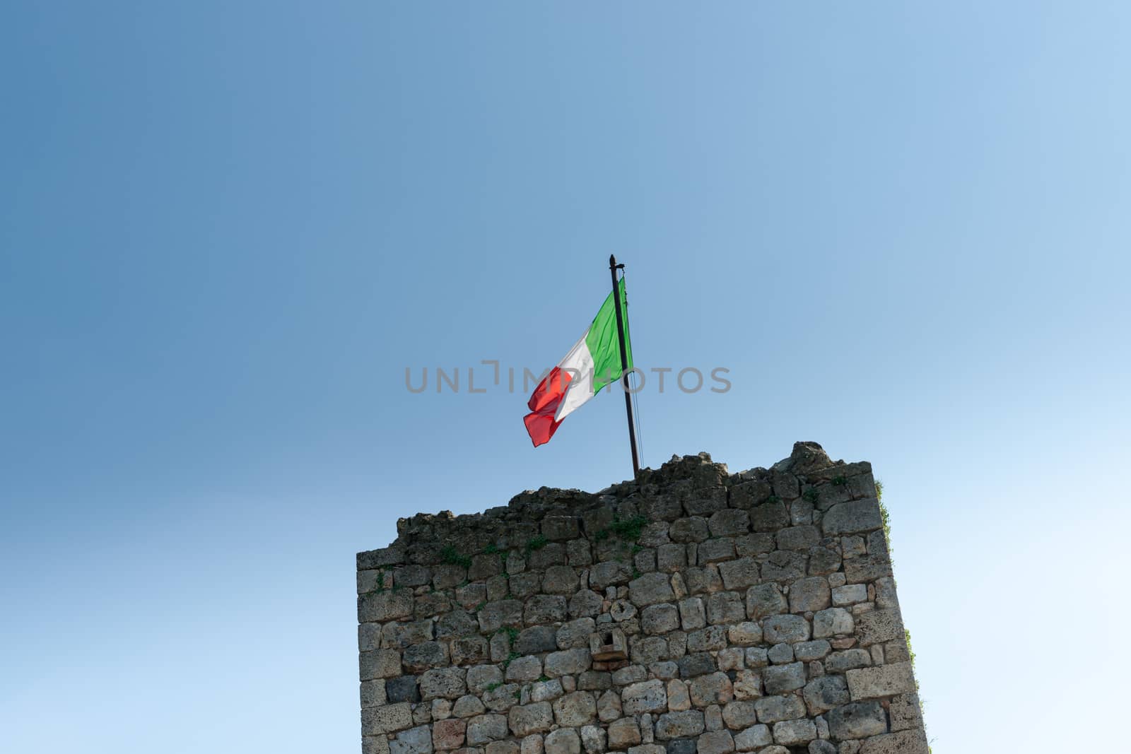 Italian flag backlit by sun above historic stone wall.