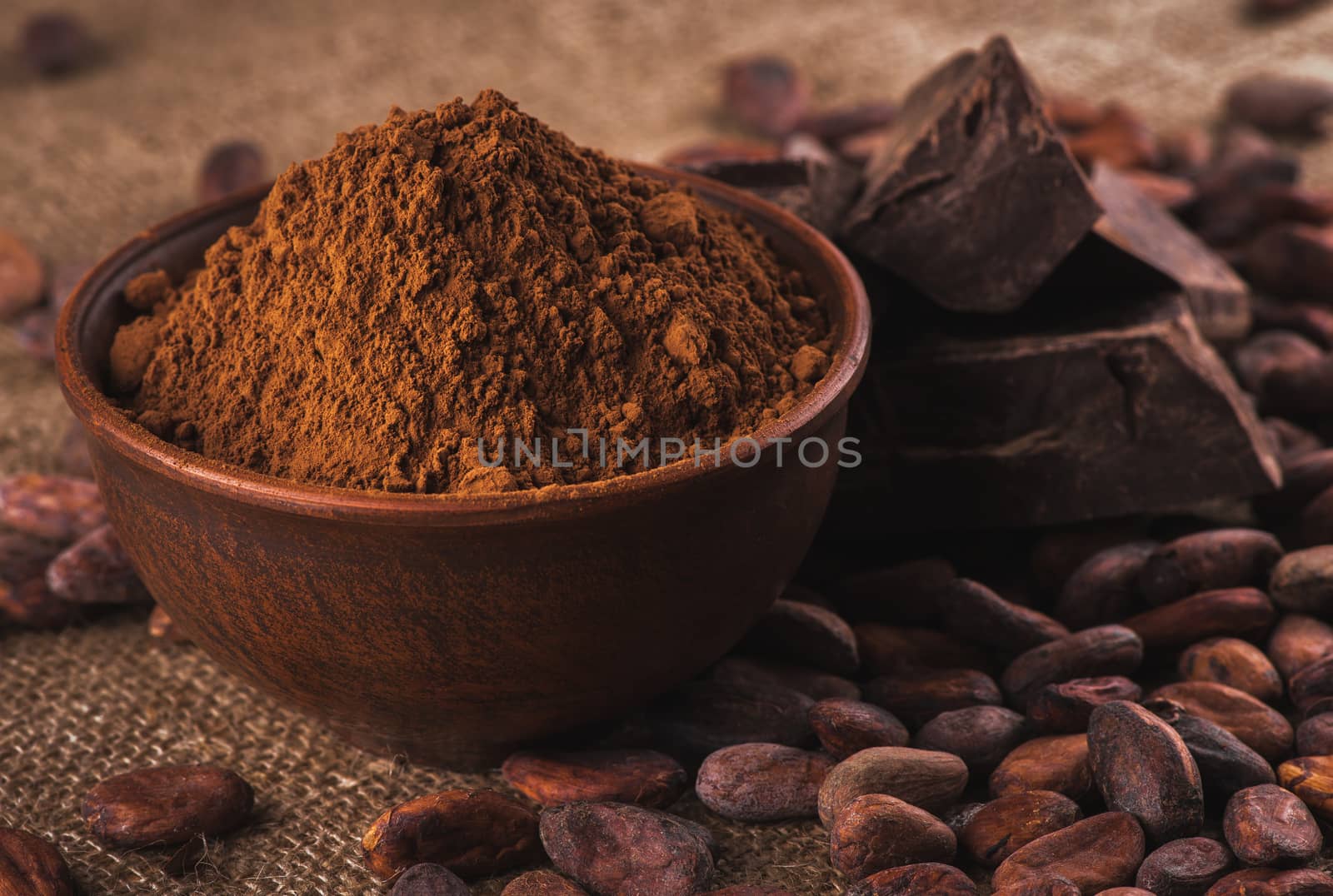crude dark cocoa powder in a brown ceramic bowl, raw cocoa beans in the peel and raw chocolate on sacking close up, ingredients for preparing chocolate and sweets
