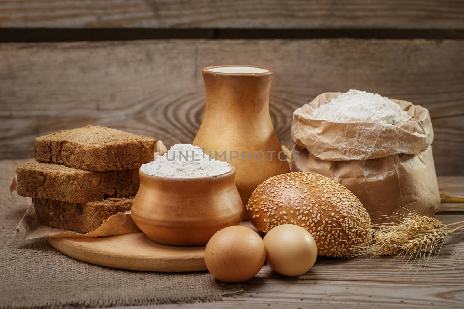 Ingredients for baking bread and pastry, milk, wheat flour, eggs, bun with sesame, rustic bread, cut into pieces, ears of wheat on the old wooden background