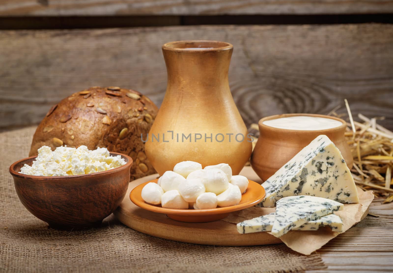 Rustic natural dairy products, whole milk, cottage cheese, cut slices of blue cheese with mold, mozzarella cheese, rustic bread, burlap, straw on the old wooden background