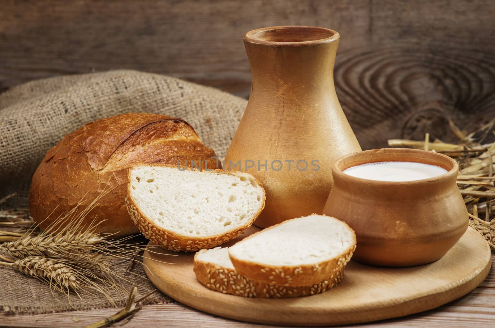 Bread and bakery products, and milk in a clay jug and bowl, stalks of wheat, burlap on the old wooden background