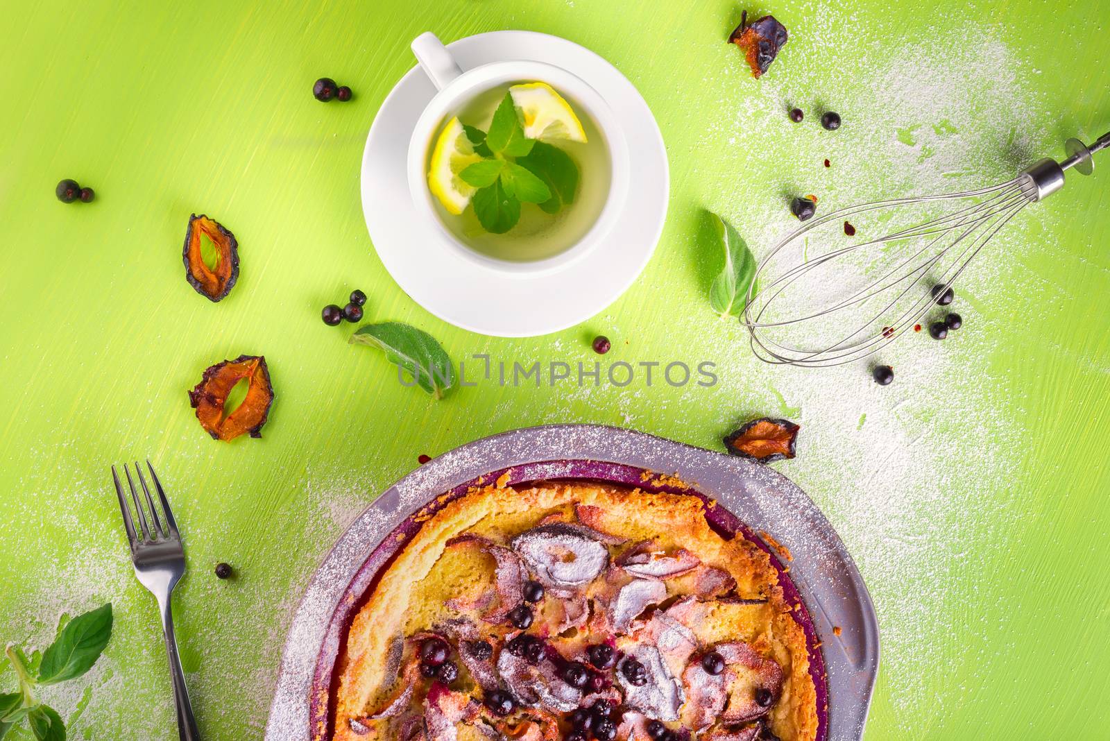 Good morning, homemade Crumble pie with black currants, plums and powdered sugar for breakfast, mint tea with lemon, fork, whisk, dried plum, mint leaves on wooden green table, top view, composition