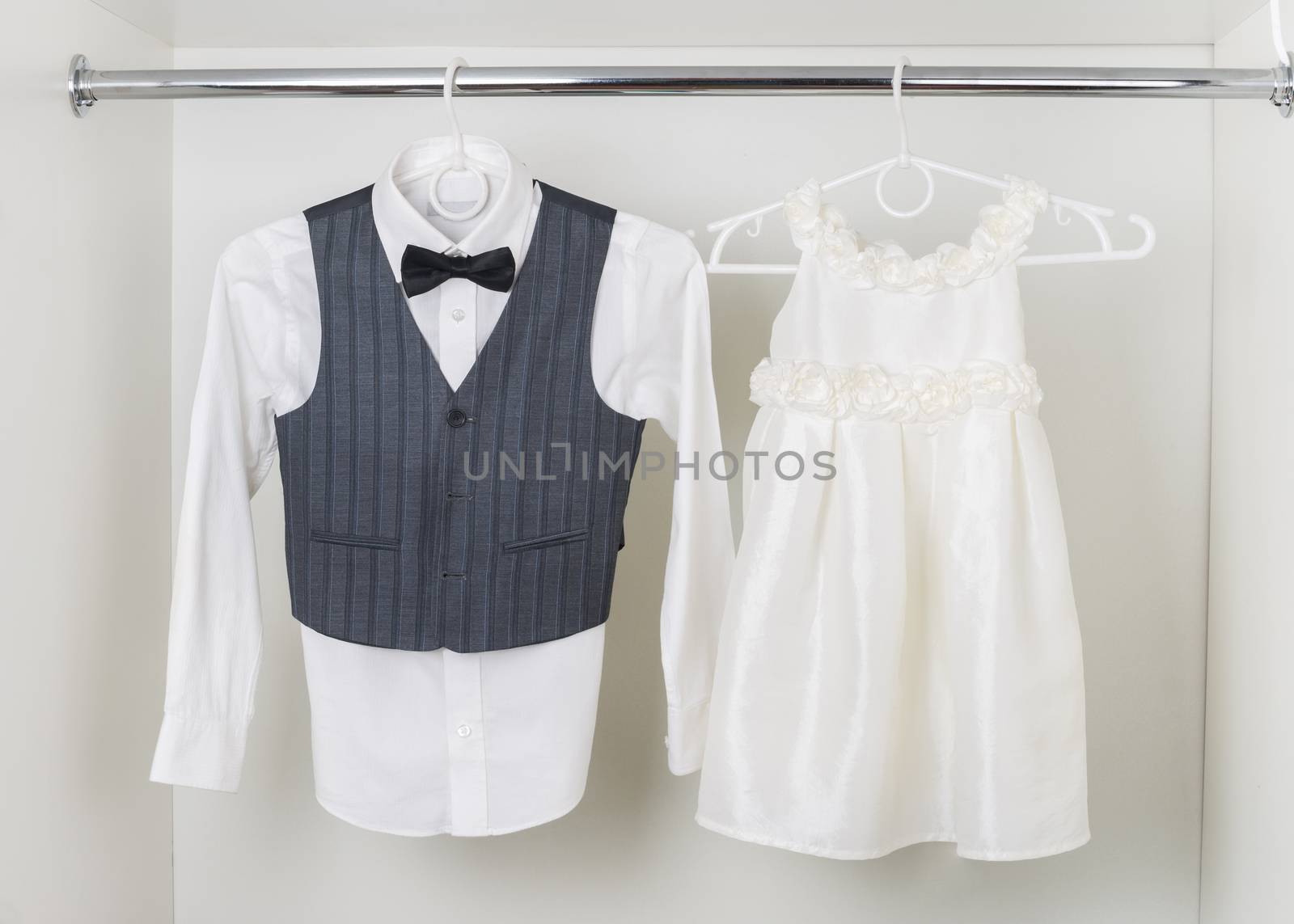 white  boy's shirt with black bow tie, vest and white elegant a little girl  dress hanging on hangers in the white wardrobe, clothes for the holiday, ceremony, wedding