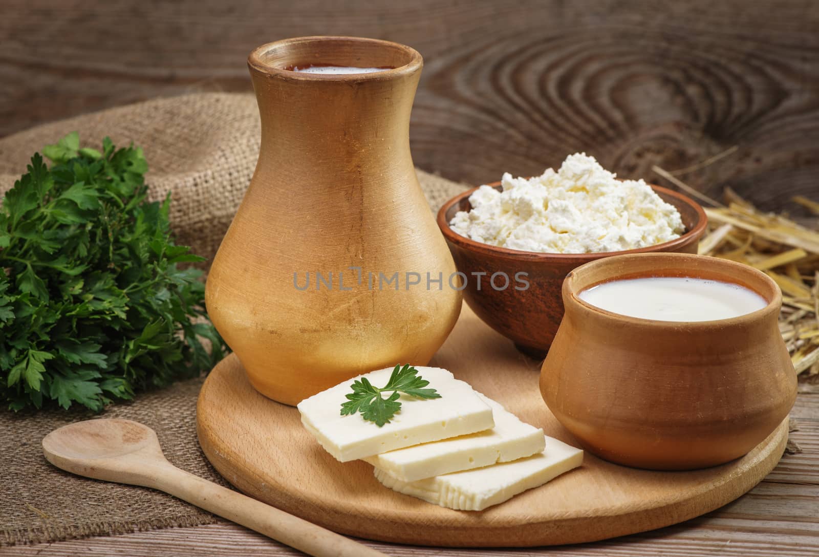 Rustic natural dairy products, whole milk in a clay jug and a cup, cottage cheese in a bowl, cut slices of cheese, parsley, burlap, straw on the old wooden background