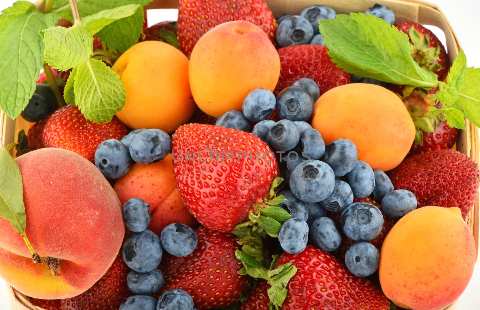 Mellow fresh summer strawberries, blueberries, apricots, peach and mint leaves in wooden basket isolated on white background, top view