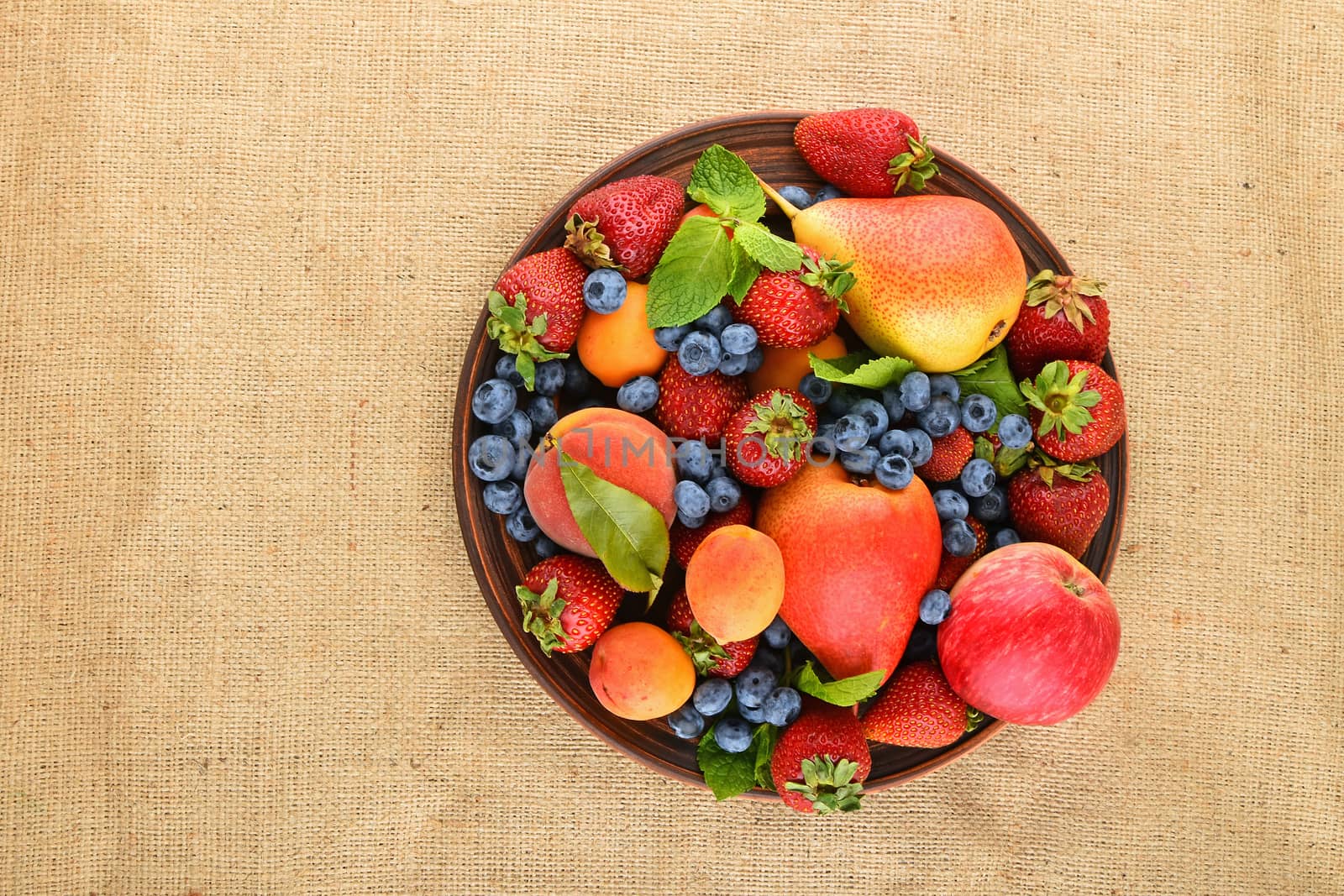Mellow fresh summer fruits and berries mix with mint leaves in ceramic plate on burlap jute canvas, strawberries, blueberries, apricots, peach, apple and pear