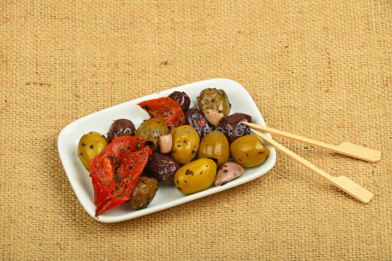 White plate of Mediterranean snack of red and green olives and pickled red pepper with skewers on burlap jute canvas