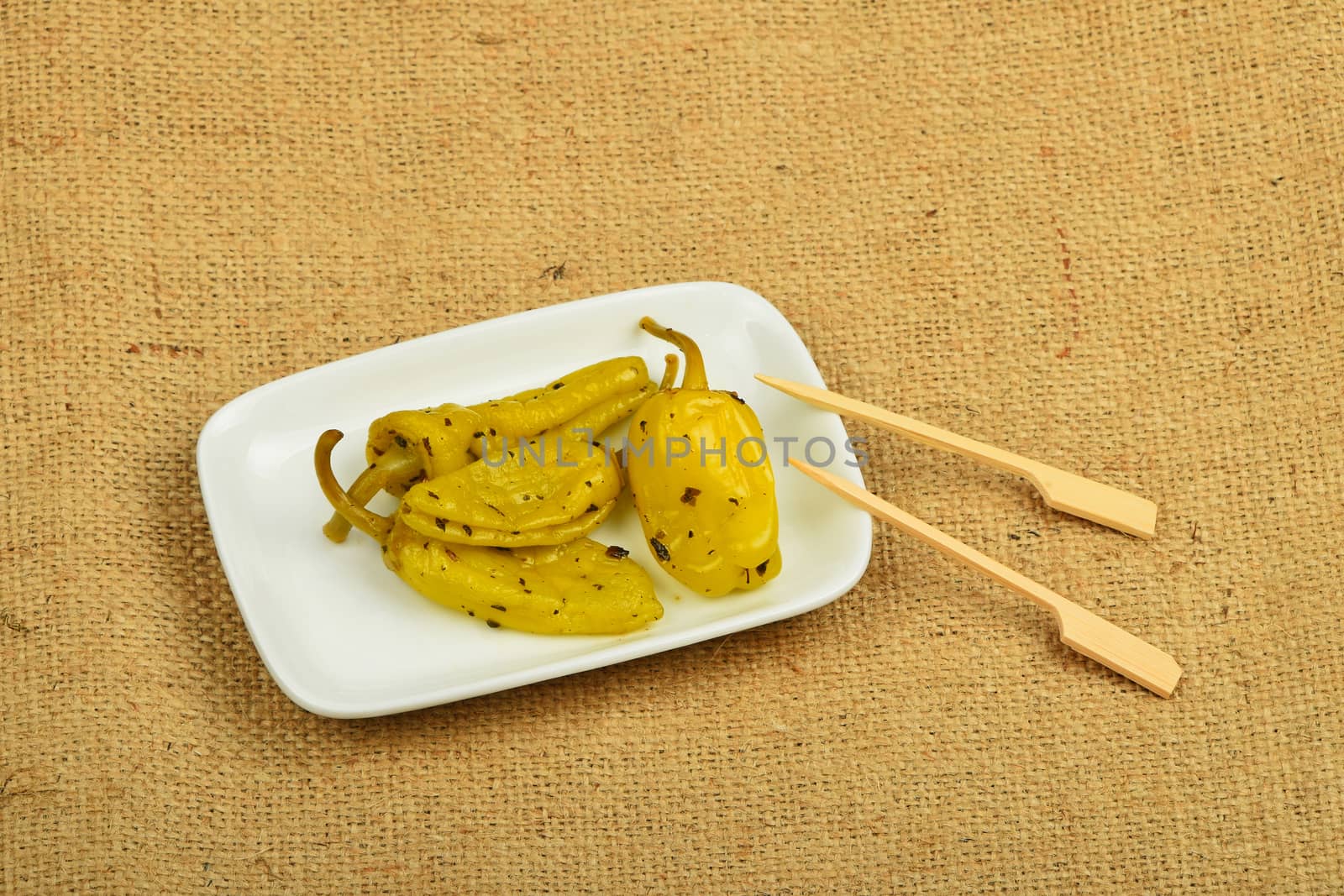 Mediterranean snack of four pickled green pepper in white plate with two skewers on burlap jute canvas